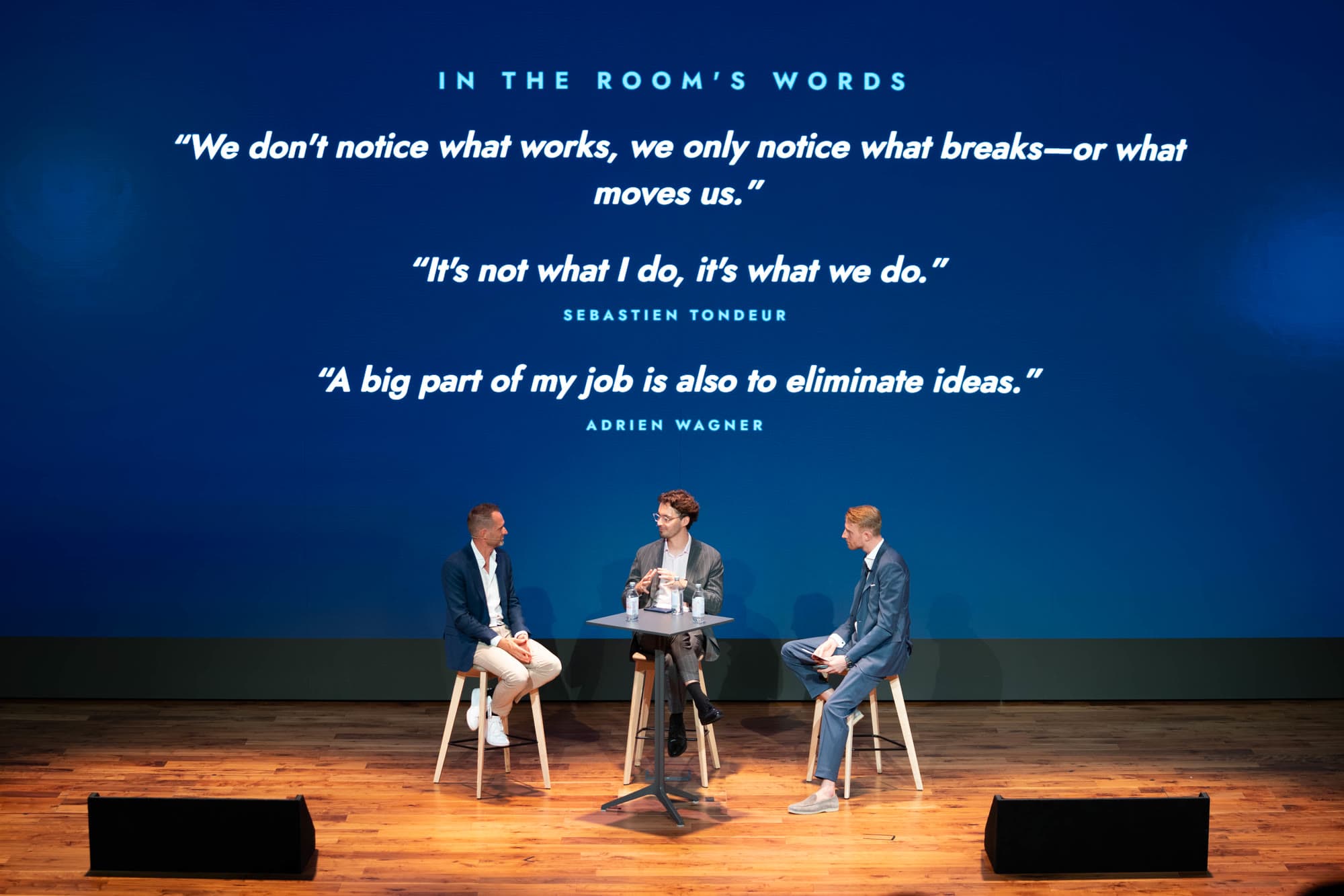Panel of three on stage under an LED wall titled In the room’s words, showing quotes from the conversation captured live by Ovee.