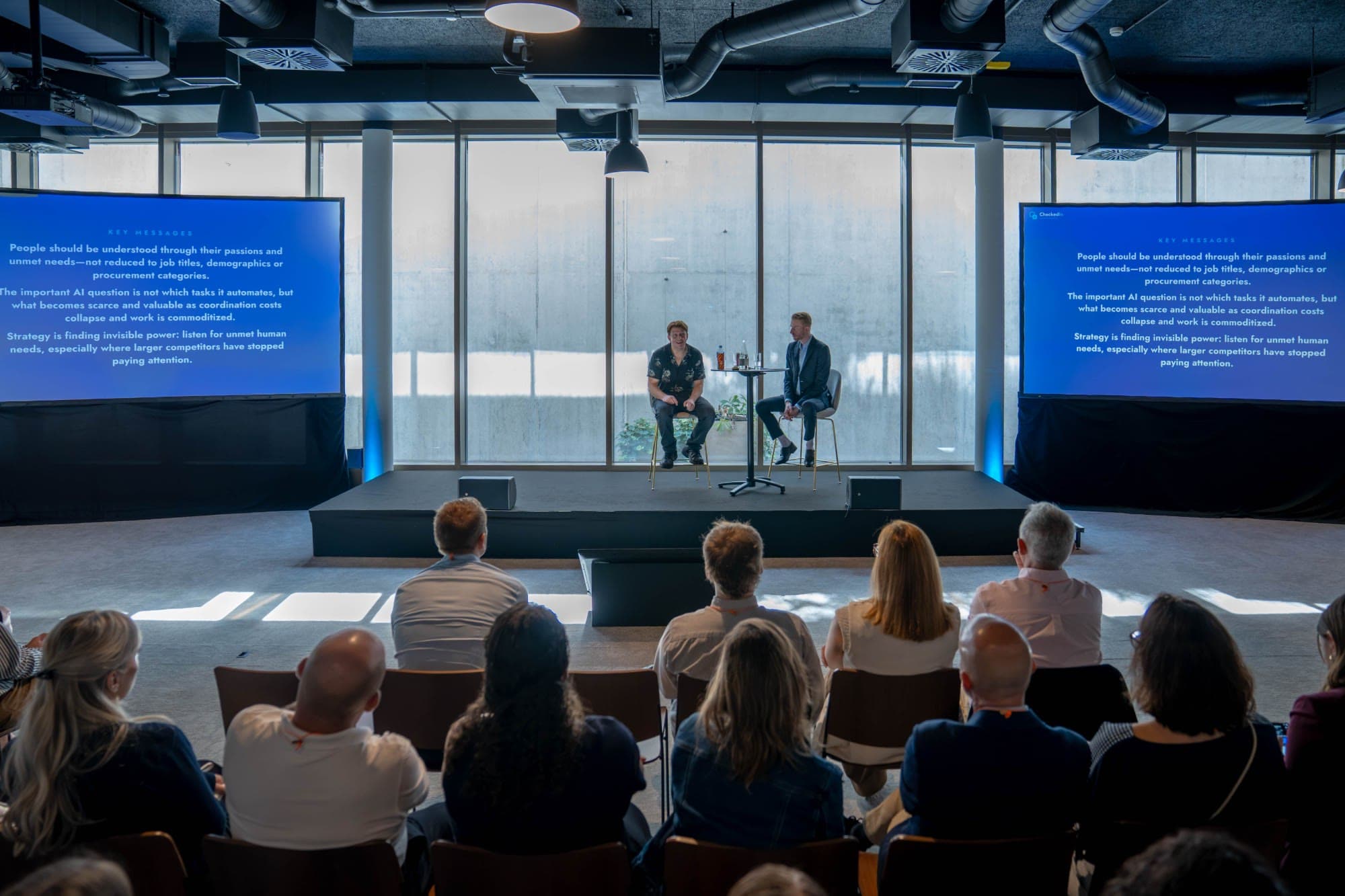 Audience facing a two-person conversation on a small stage, flanked by two screens showing key messages from the talk.