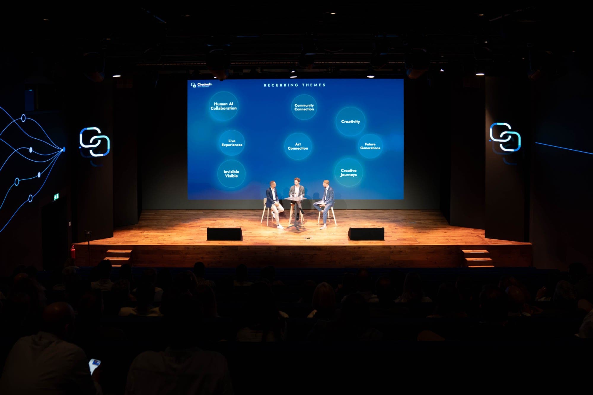Panel of three on a lit stage in front of a large LED wall showing eight recurring themes generated during the session.