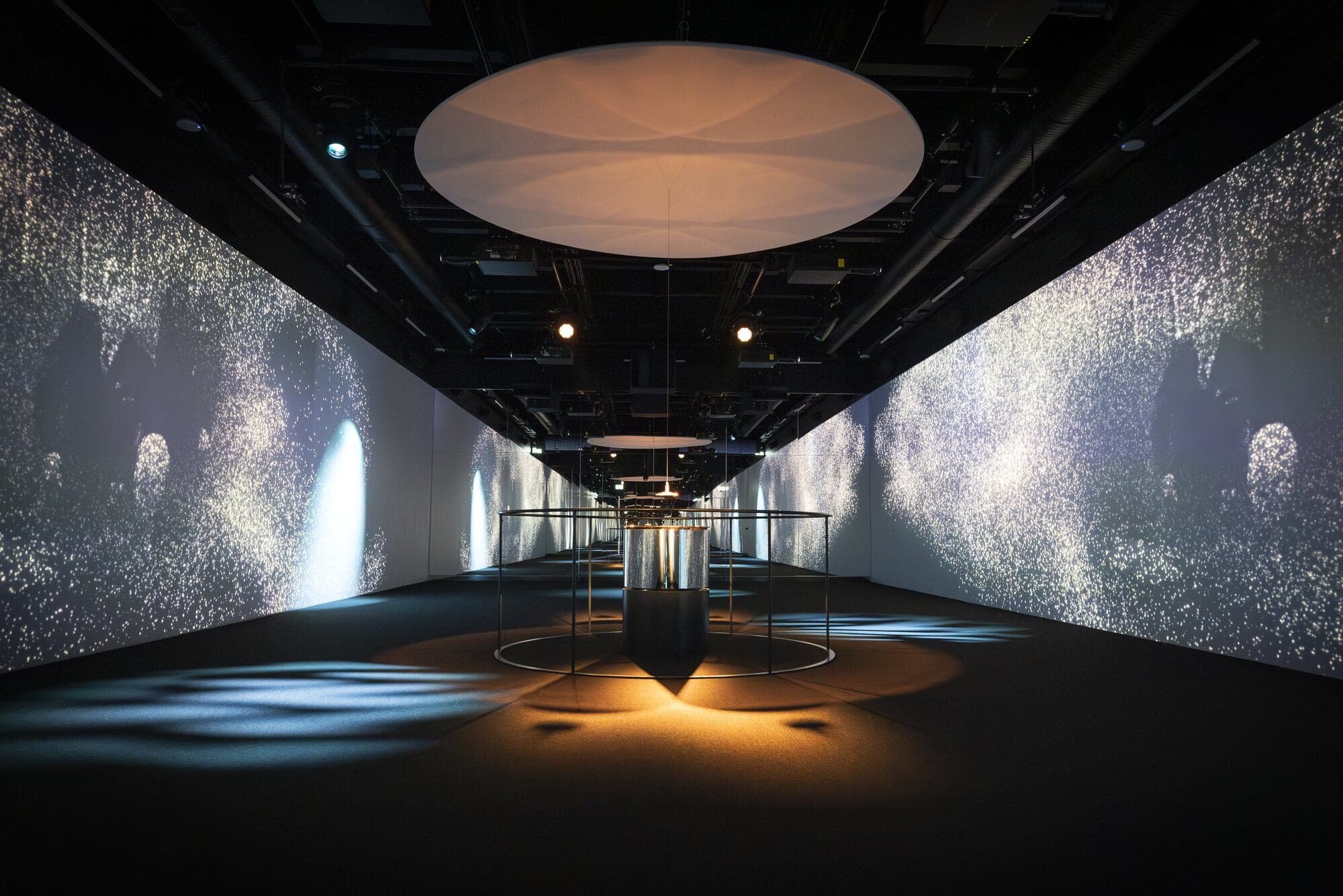 A long dark pavilion with projected walls and a central lit totem under a circular ceiling light.
